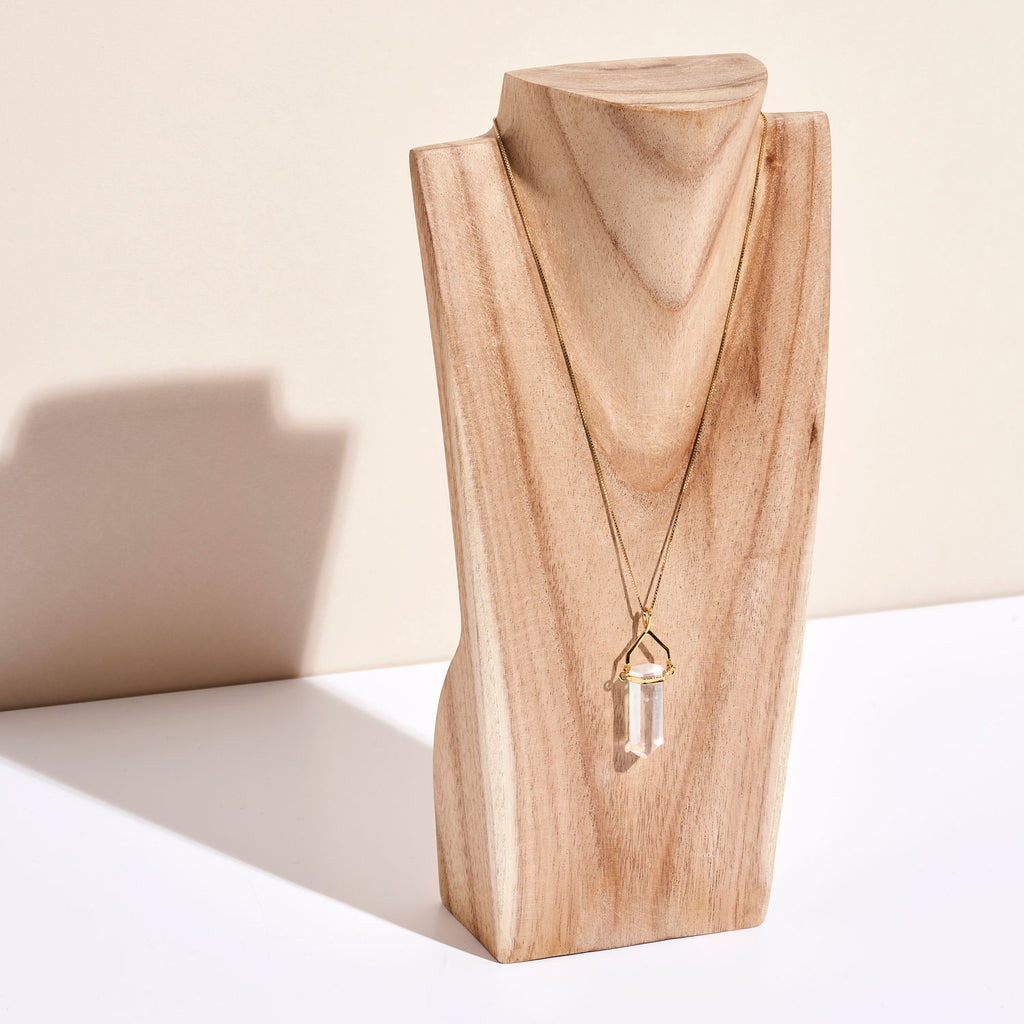 Photo d'un pendentifs en pierre de Cristal de roche en pointe orné d'un fermoir et d'une chaine dorés de la marque Let's Play On The Moon sur un portant en bois